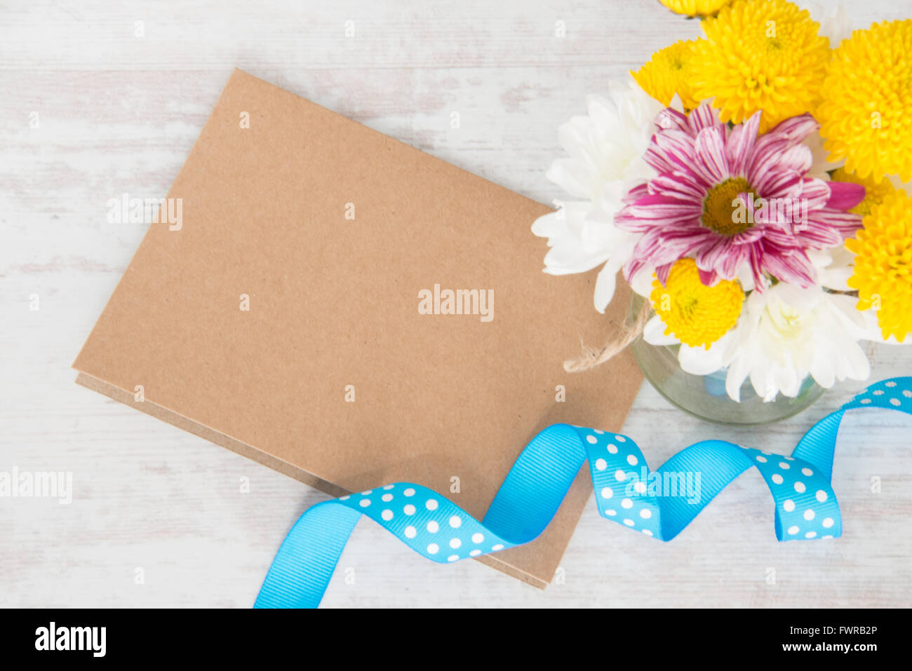 Spring flowers bouquet in a jar vase with kraft card note, envelope and blue dotted ribbon on a white wood rustic background Stock Photo