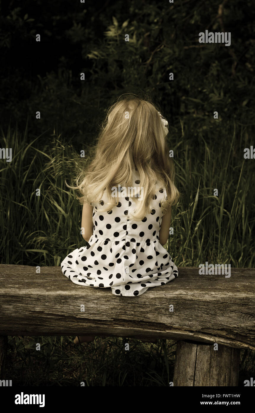 back view of blond girl in dotted dress sitting on bench Stock Photo