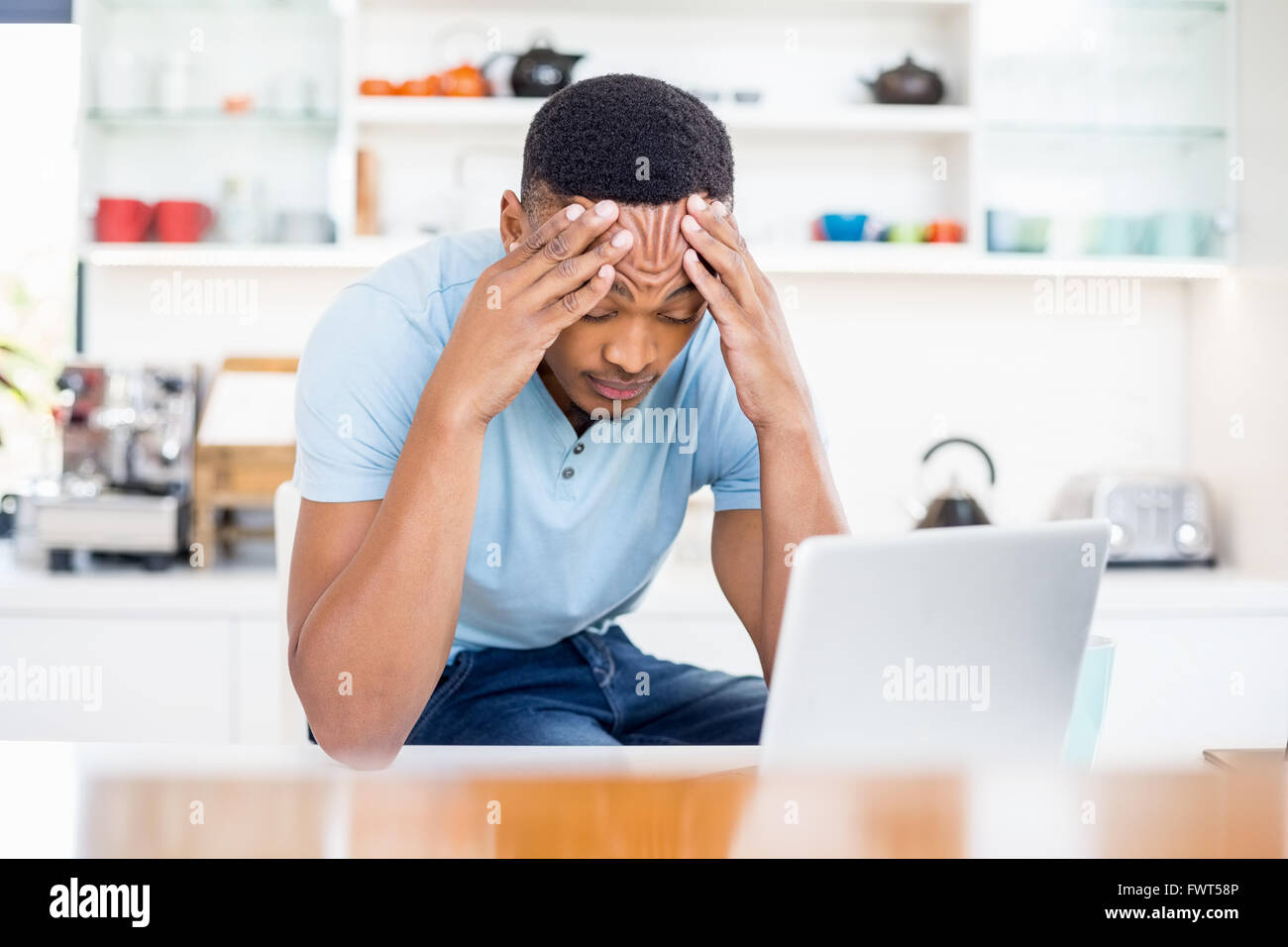 Tense young man sitting in kitchen with laptop Stock Photo