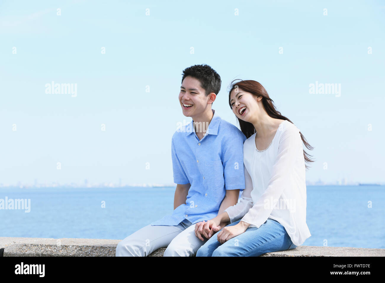 Young Japanese couple sitting on concrete wall by the sea Stock Photo ...
