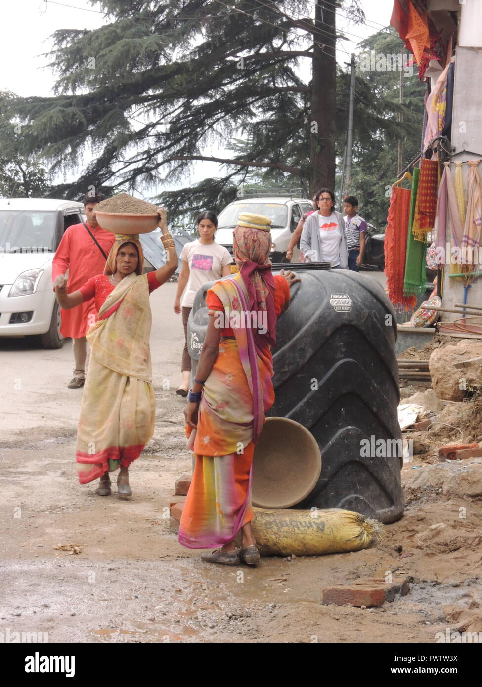 Poor Indian woman carries rubble on her head working at a construction site while modern cars go by in the background Stock Photo