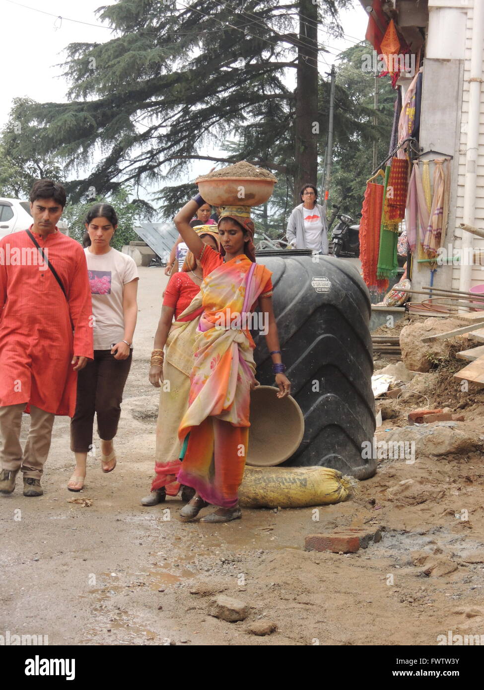 Poor Indian woman carries rubble on her head working at a construction site, a wealthy Indian woman in western dress walks past Stock Photo