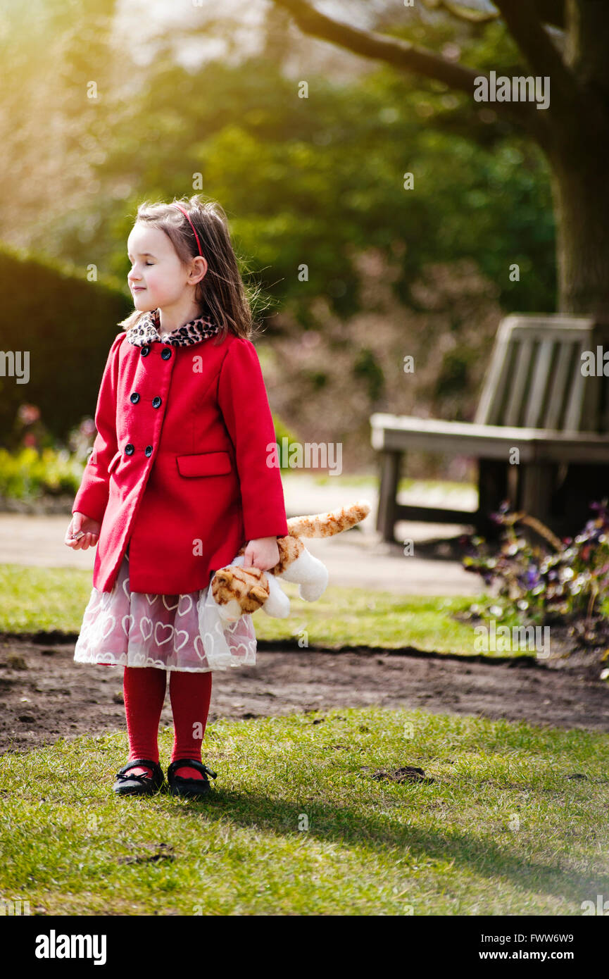 a lost small girl wearing red coat standing in park holding a toy Stock Photo