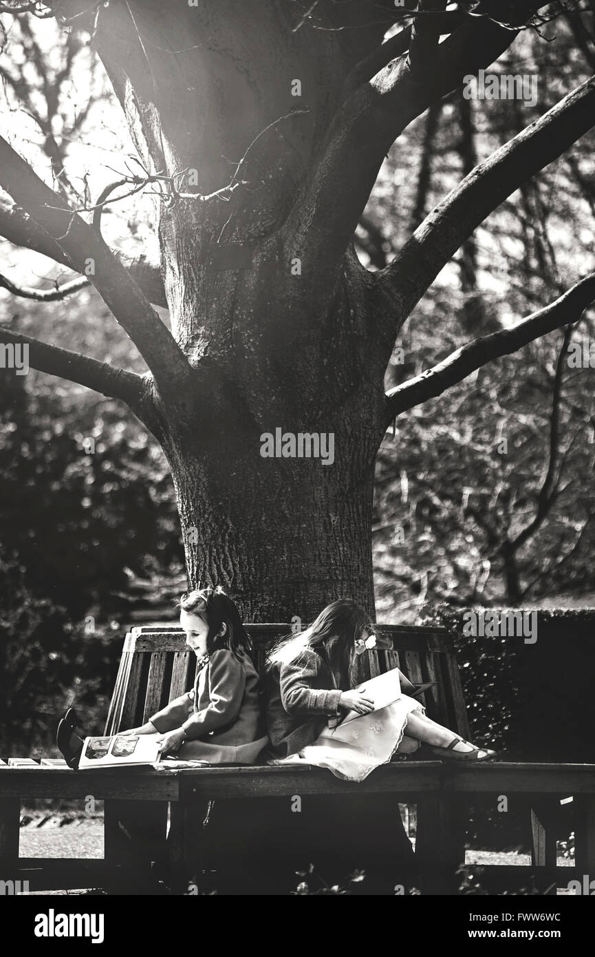two little girls sisters sitting on bench under tree and read books Stock Photo
