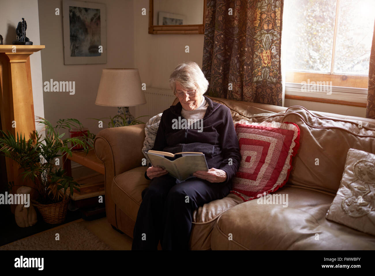 Senior Woman Sitting On Sofa Reading Book At Home Stock Photo - Alamy