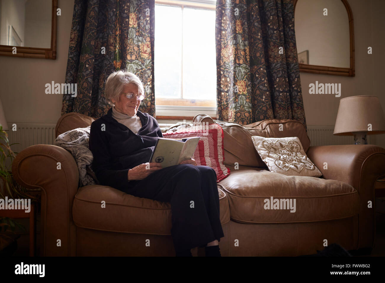Senior Woman Sitting On Sofa Reading Book At Home Stock Photo - Alamy