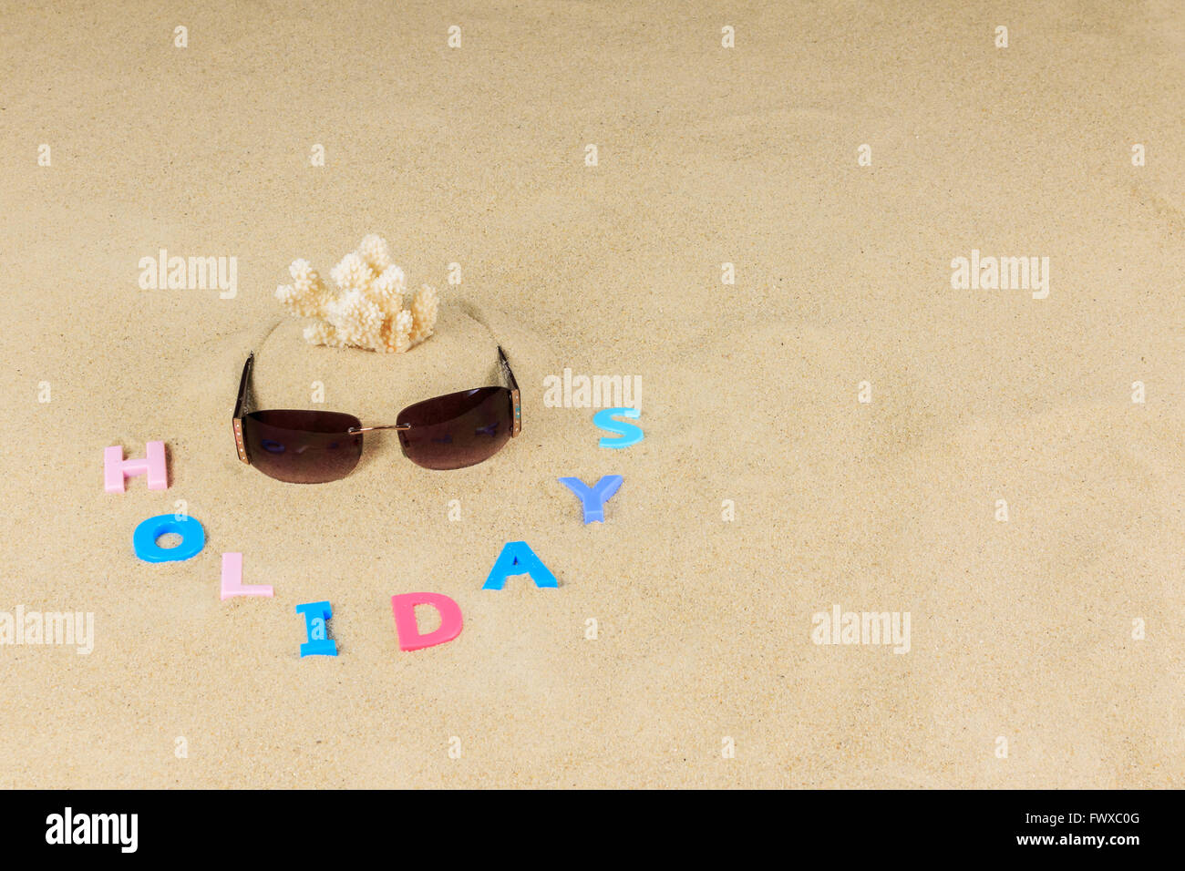 Pair of  sunglasses, pieces coral and coloured letters spelling holidays on a sand background Stock Photo