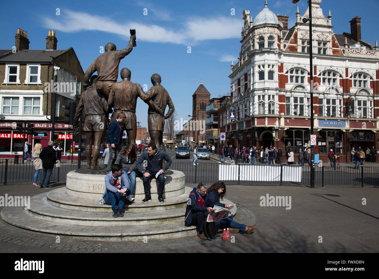 Fans sitting at the World Cup statue on Barking Road, with the Boleyn pub, a regular gathering place for home fans, in the background before West Ham United hosted Crystal Palace in a Barclays Premier League match at the Boleyn Ground. The Boleyn Ground at Upton Park was the club's home ground from 1904 until the end of the 2015-16 season when they moved into the Olympic Stadium, built for the 2012 London games, at nearby Stratford. The match ended in a 2-2 draw, watched by a near-capacity crowd of 34,857. Stock Photo