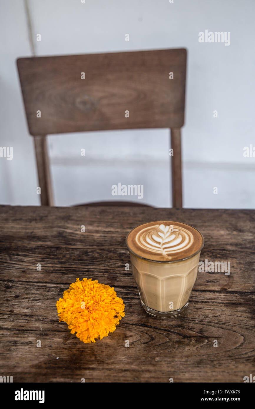 Cafe latte in glass with latte art and orange flower on wooden table and chair in background Stock Photo