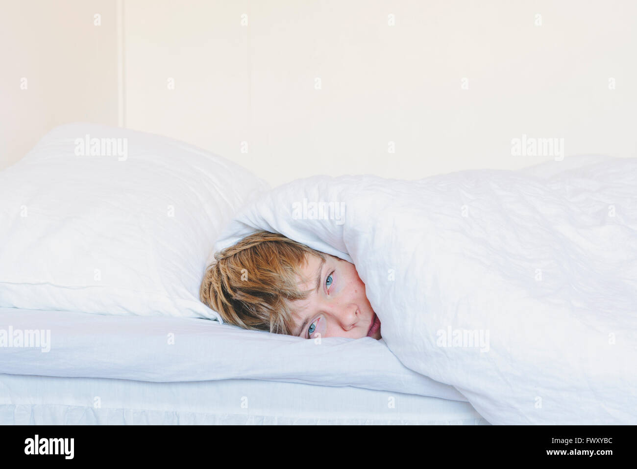 Finland, Helsinki, Portrait of young man lying in bed under white blanket Stock Photo