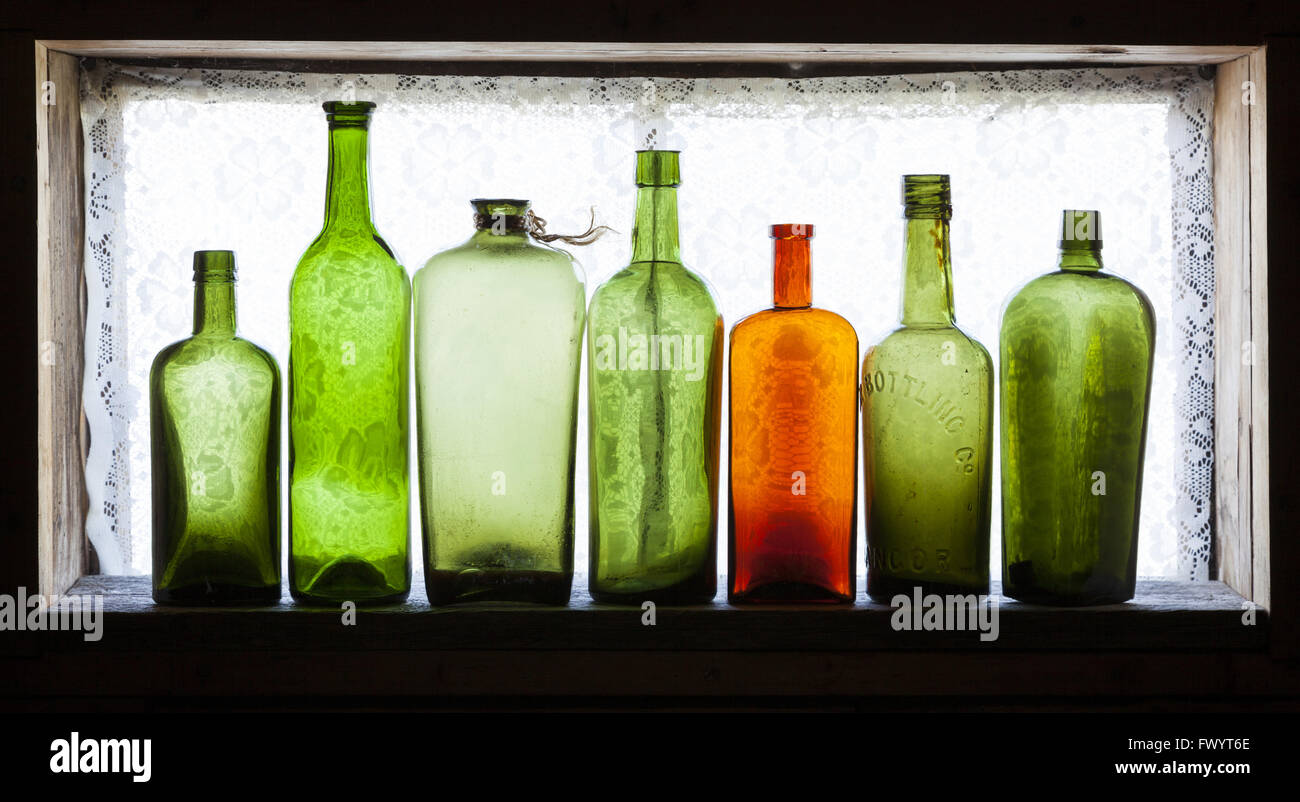 Orange bottle among green antique bottles on old windowsill against daylight. Stock Photo
