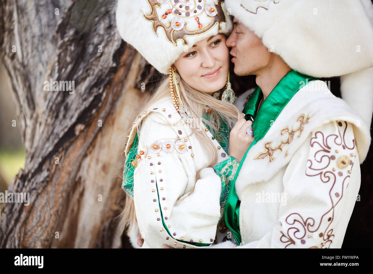 Couple in ethnic costumes embrace on background of textured wood, groom kisses bride at cheek. Stock Photo