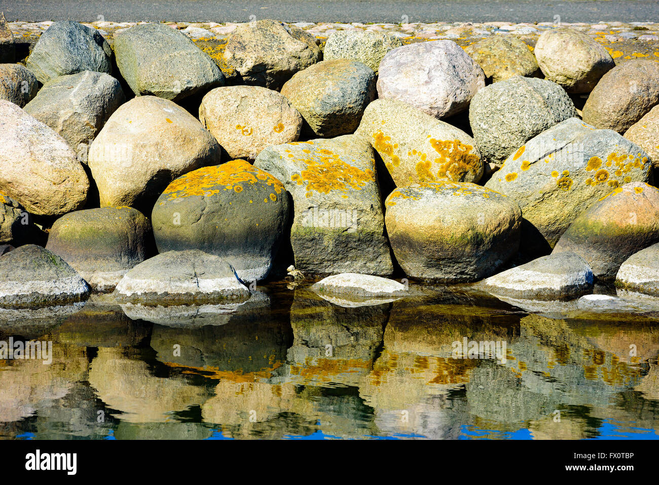 Close up of a stone pier with lichen covered granite boulders. Seawater with fine reflections in lower part and some pavement vi Stock Photo