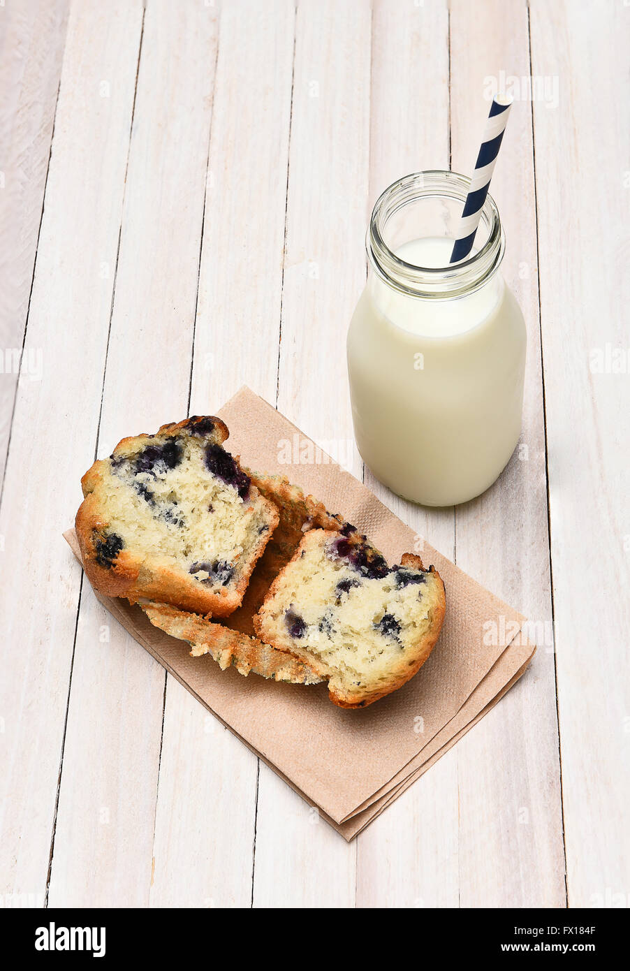 High angle view of a blueberry muffin and bottle of milk on a rustic white table. The muffin is broken in half on a napkin. Vert Stock Photo