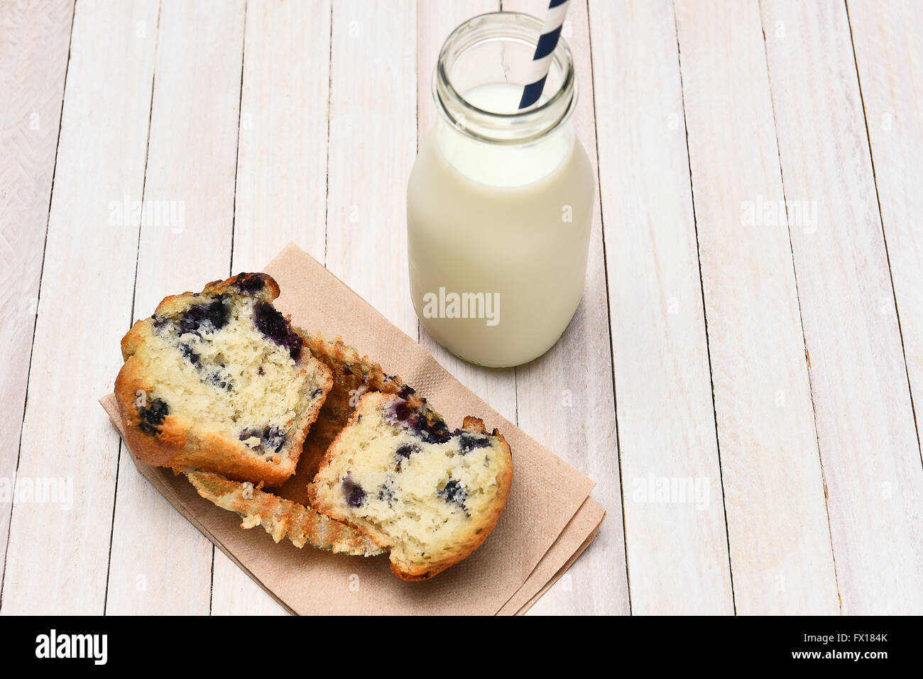 High angle view of a blueberry muffin and bottle of milk on a rustic white table. The muffin is broken in half on a napkin. Stock Photo