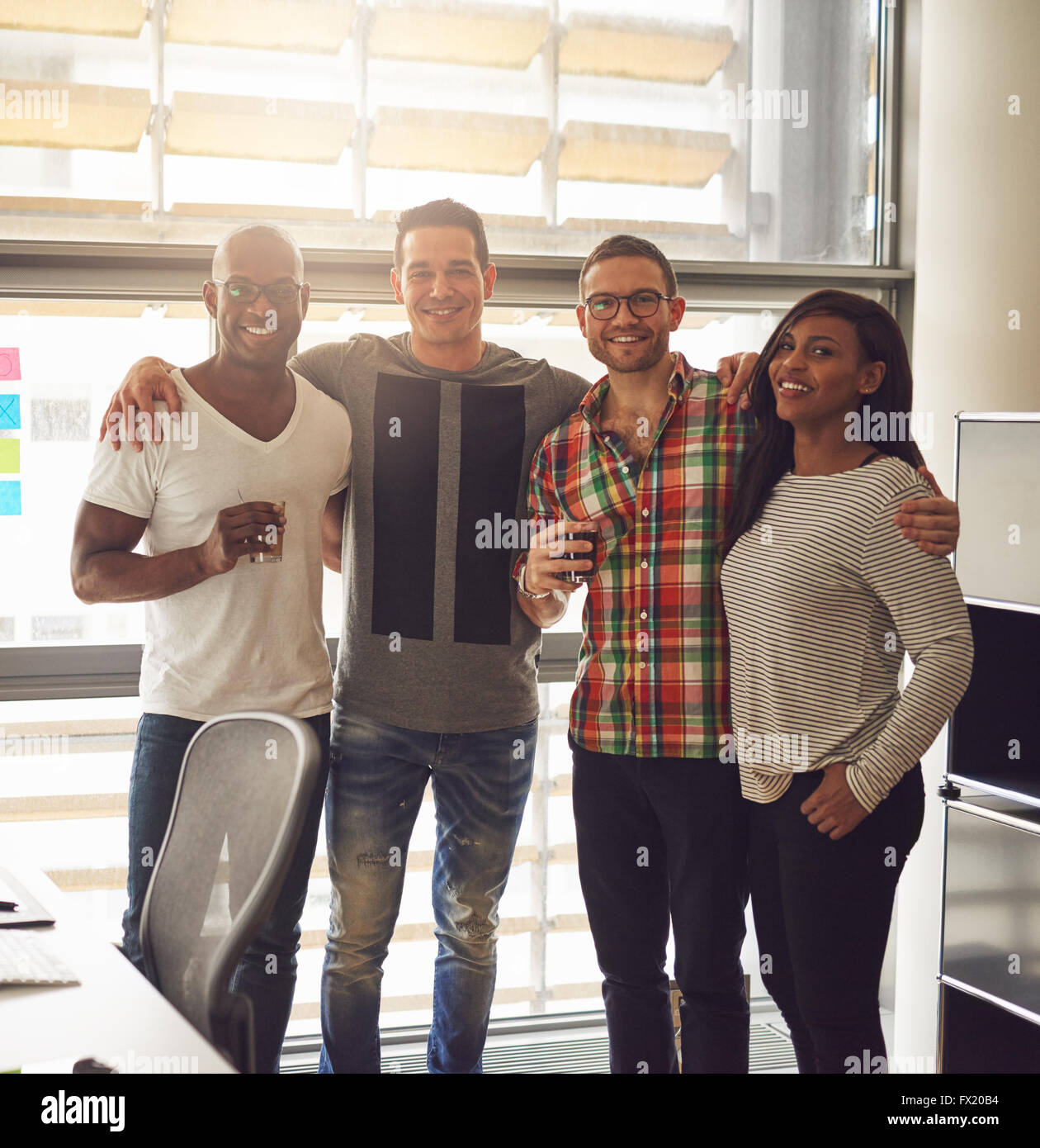 Group of four diverse friends dressed in casual clothing and holding drinks while standing close to each other in office with la Stock Photo