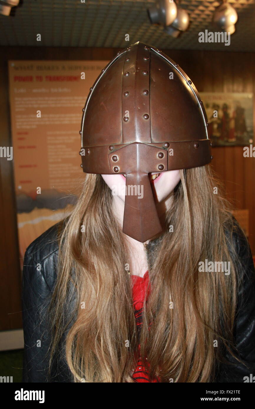 Head and shoulders image of a smiling girl with long dark blond or light brown hair wearing a too big bronze Viking helmet Stock Photo