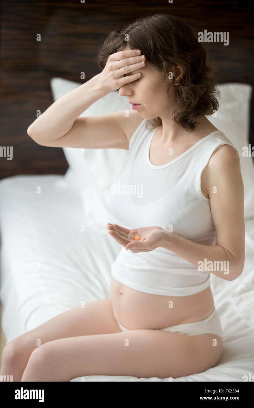 Health care, treatment, supplements. Young pregnant woman sitting indoor on the bed holding round orange pill in her palm Stock Photo
