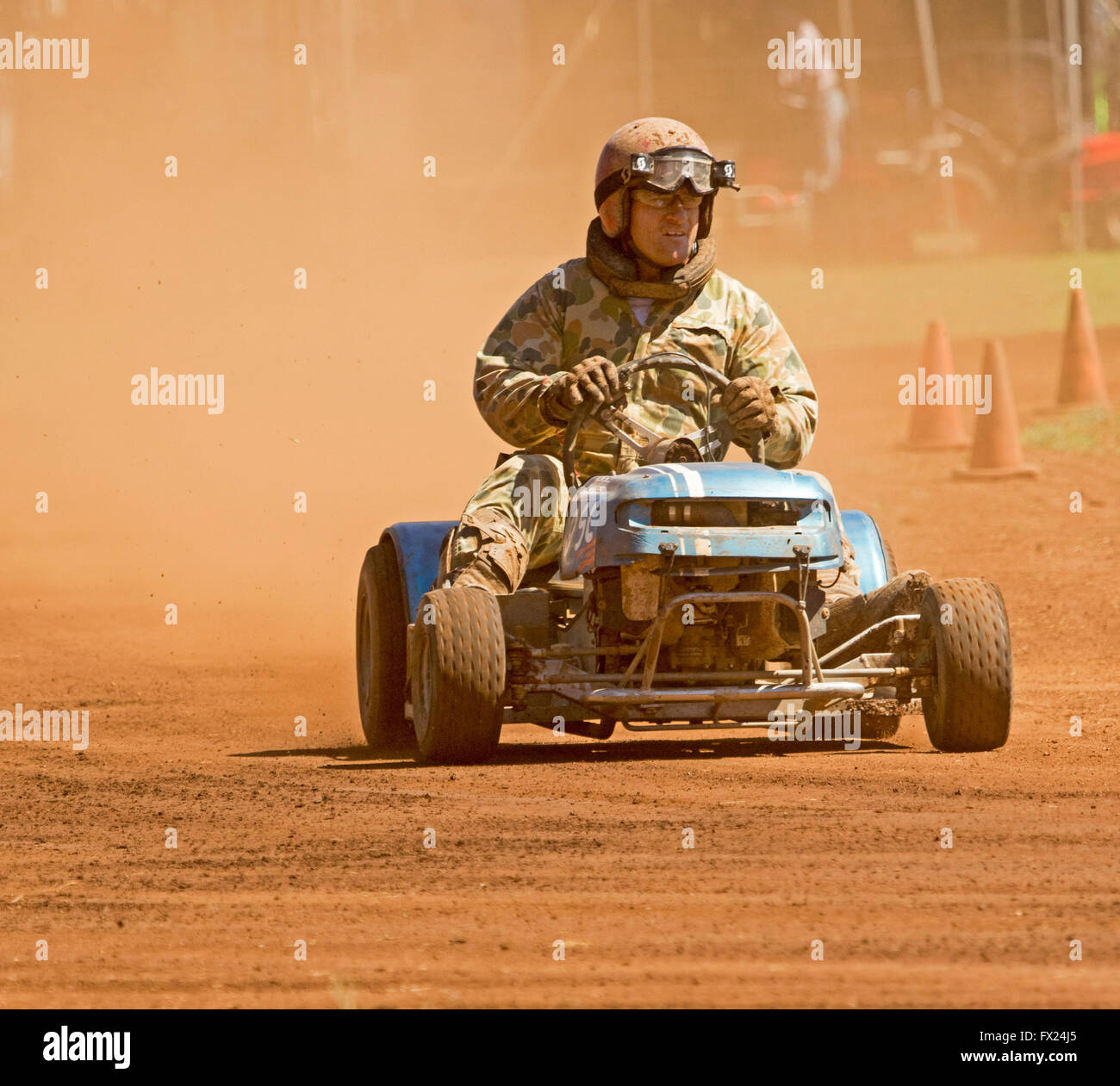 Man with helmet & overalls driving motor mower emerging from cloud of red dust on track in unusual Australian motor racing sport Stock Photo