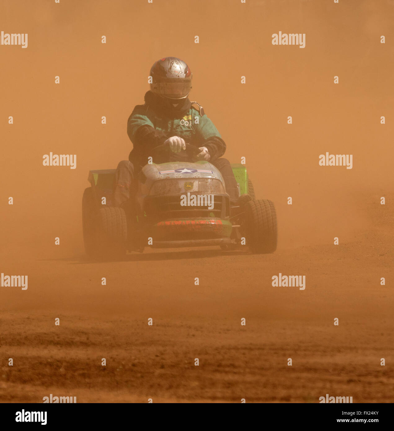 Man with helmet & overalls driving motor mower emerging from cloud of red dust on track in unusual Australian motor racing sport Stock Photo