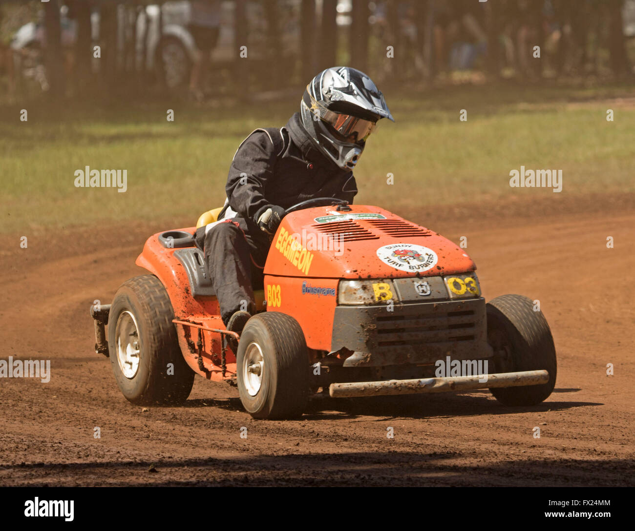 Man, dark helmet & overalls, driving bright orange Husqvarna motor mower on dusty track in unusual Australian motor racing sport Stock Photo