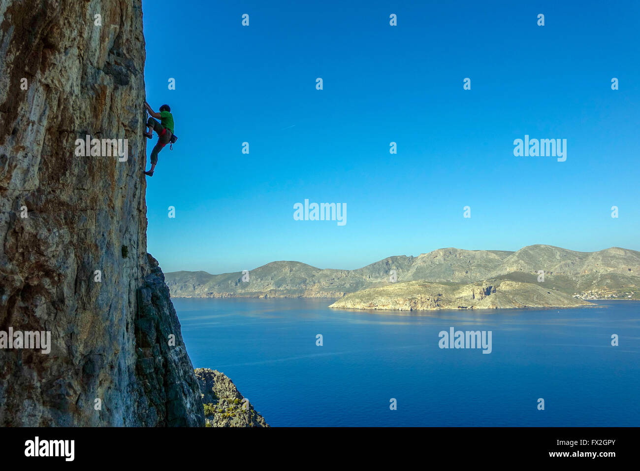 small rock climber silhouetted on steep cliff with blue sea behind Stock Photo