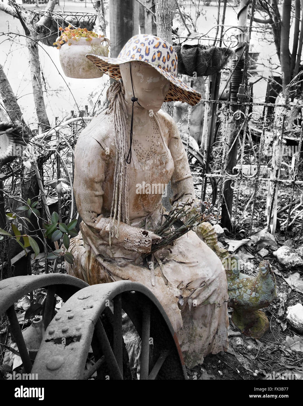 An old crumbling but beautiful statue of a woman with flowers and a cheetah print brimmed hat in a weedy garden, partially desat Stock Photo