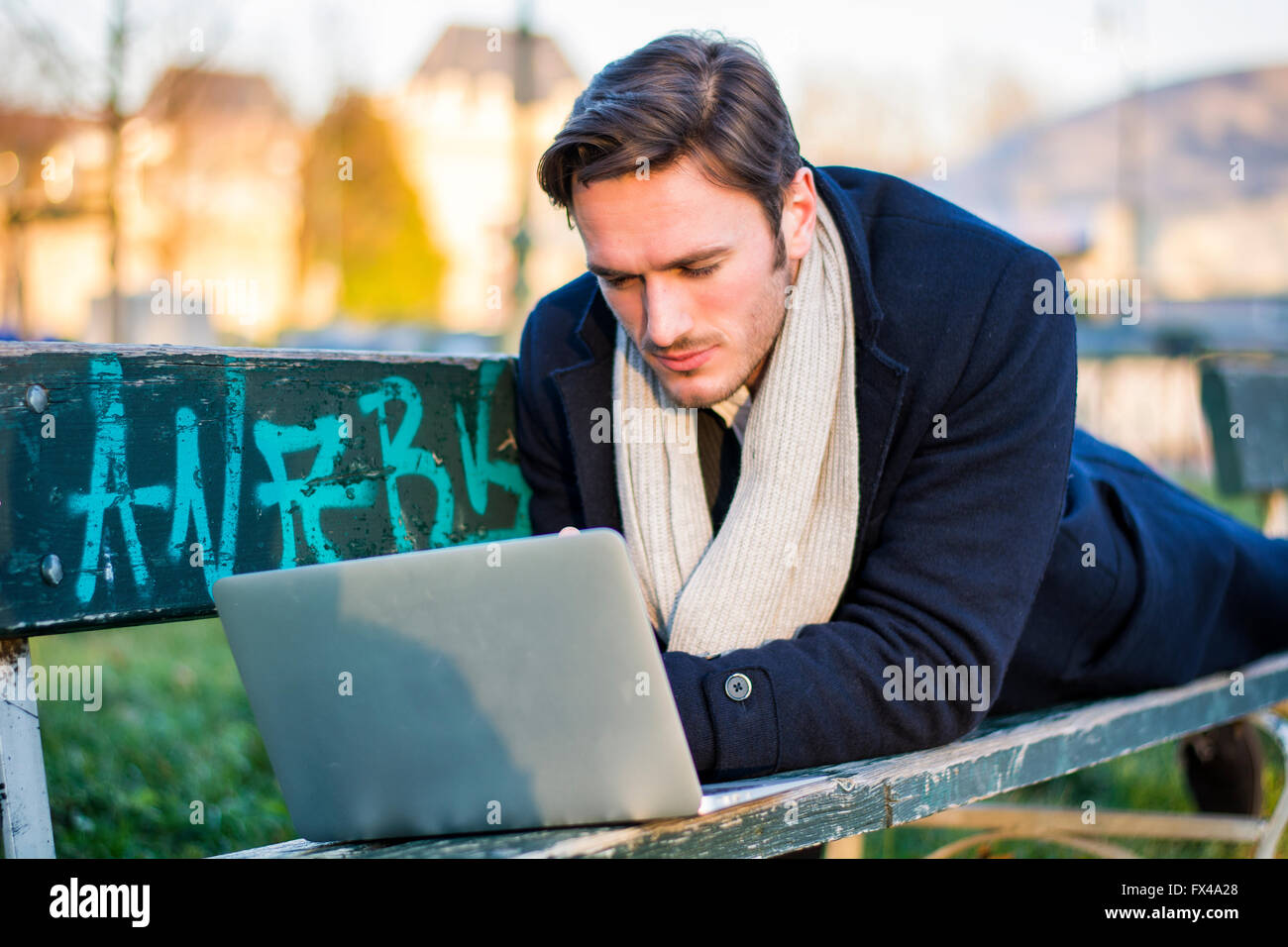 Handsome elegant businessman sitting on a wooden bench working outdoors in an urban park typing information onto his laptop comp Stock Photo