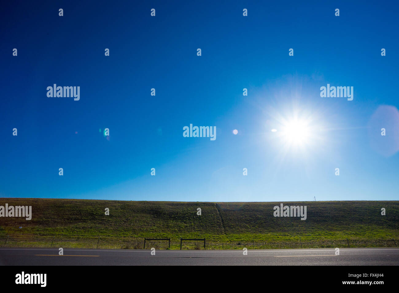 The bright sun in the blue sky causes this great sun flare along a dam and a road for a unique abstract nature sky landscape in Stock Photo