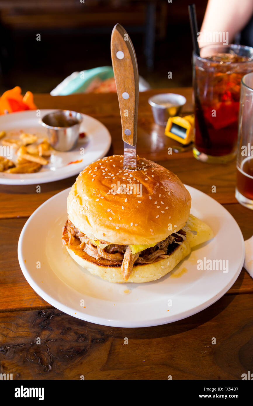 This sandwich or burger on a sesame bun has a knife in the top of it and has pulled pork and cuban spices and sauce on it. Stock Photo
