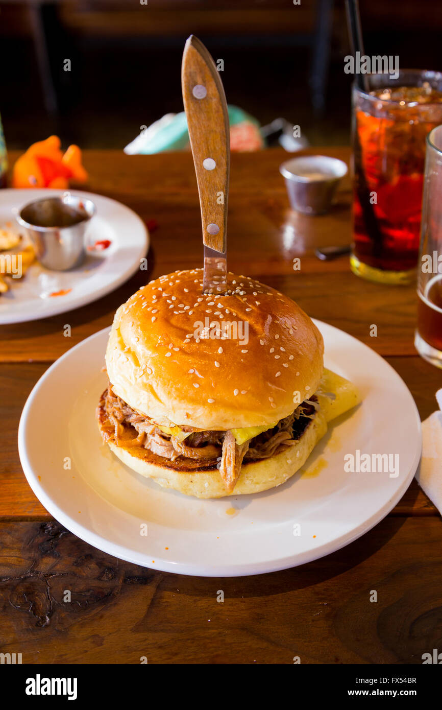 This sandwich or burger on a sesame bun has a knife in the top of it and has pulled pork and cuban spices and sauce on it. Stock Photo