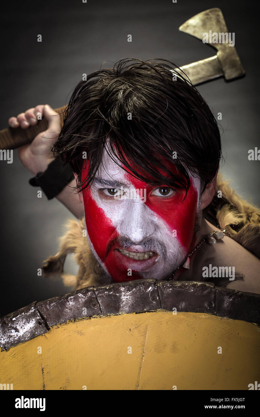 savage warrior with war painted face, hiding behind the shield of combat and furiosly swings axe Stock Photo