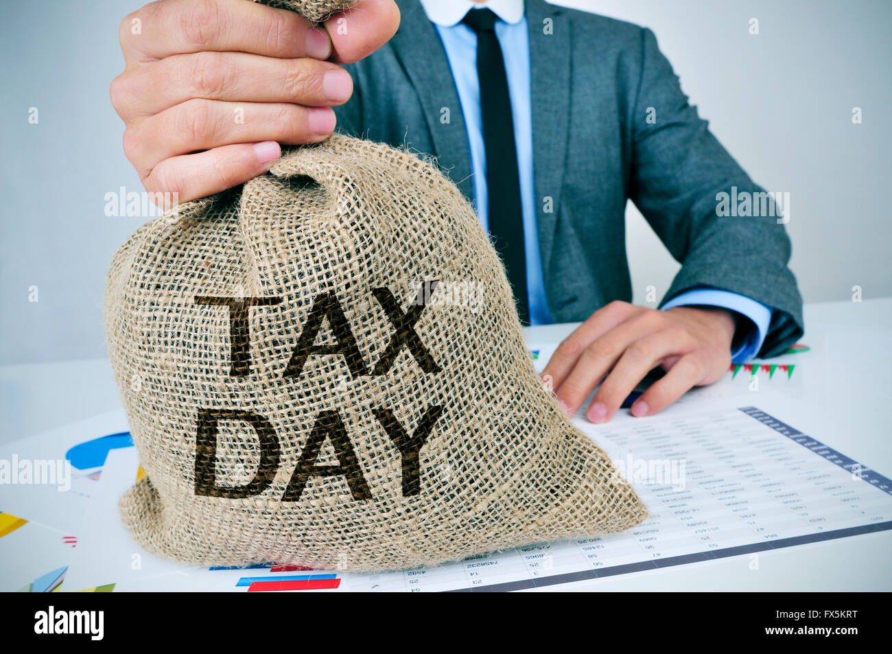 closeup of a young caucasian man sitting at his office desk holding a burlap money bag with the text tax day written in it Stock Photo