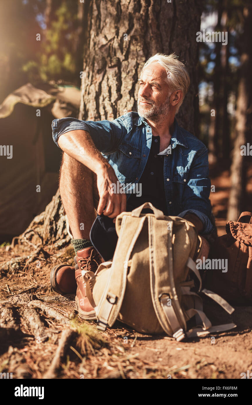 Portrait of senior man looking away while sitting by a tree. Senior man with a backpack outdoors on a summer day. Stock Photo