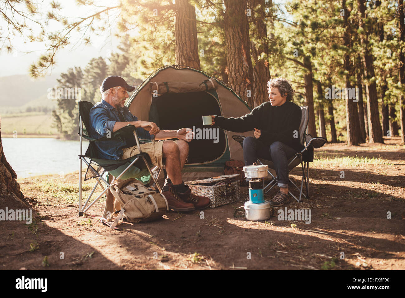 Portrait of senior couple camping by a lake, with woman giving a cup of coffee to man. Stock Photo
