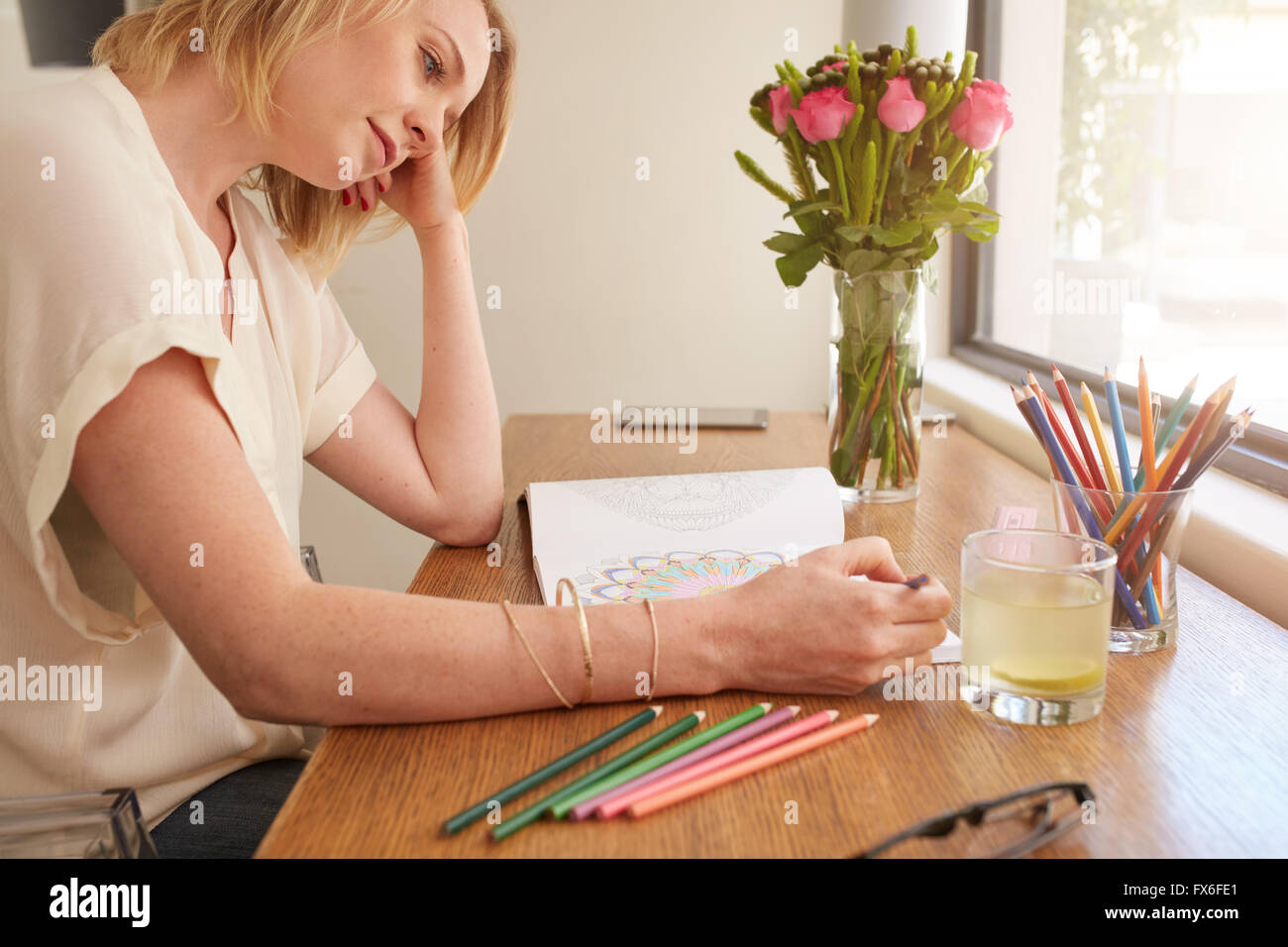 Woman drawing an adult coloring book while comfortably sitting at table by a window. Stock Photo
