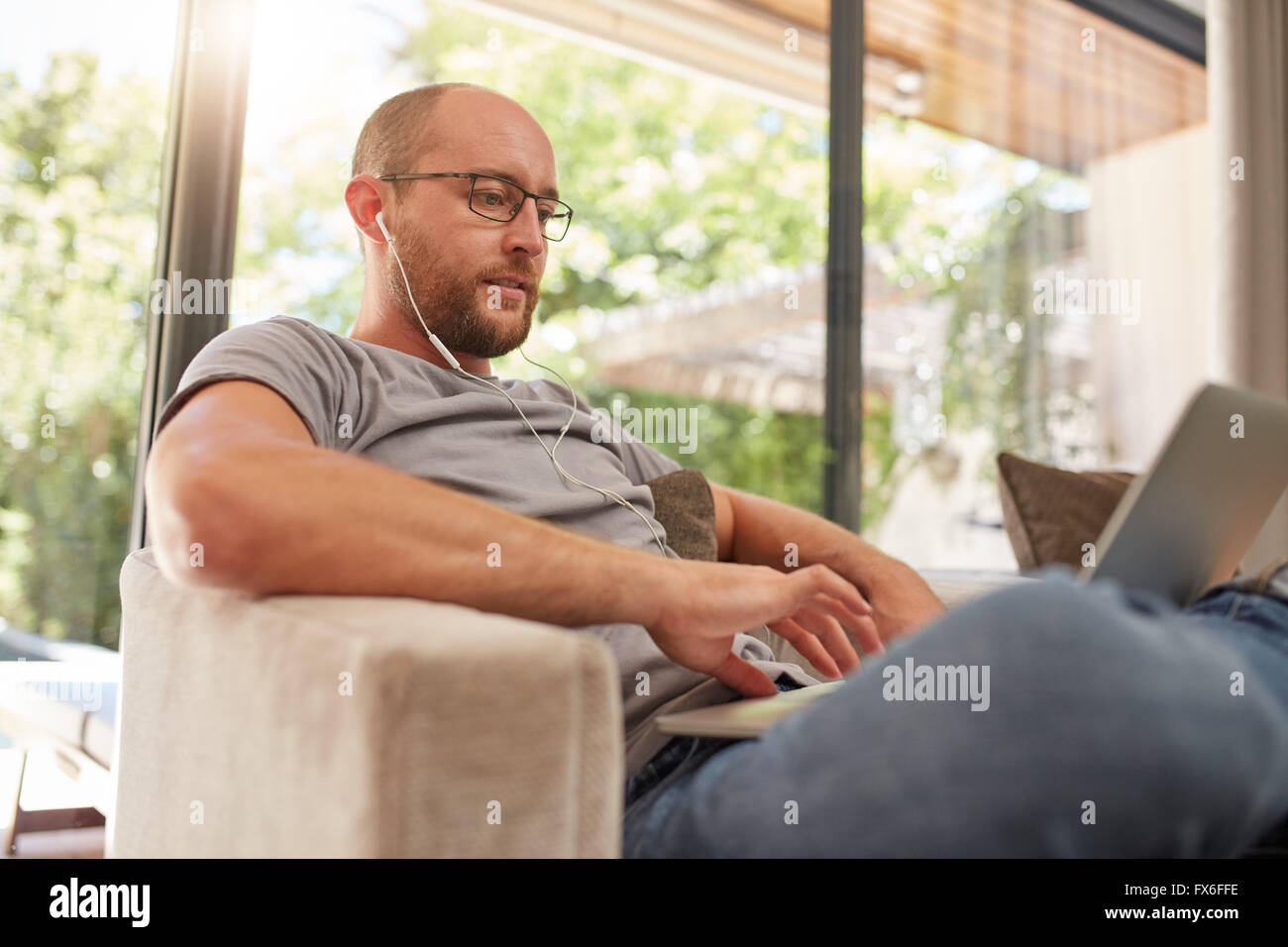 Handsome mature man using his laptop while relaxing at home, he is with earphones sitting on sofa at home. Stock Photo