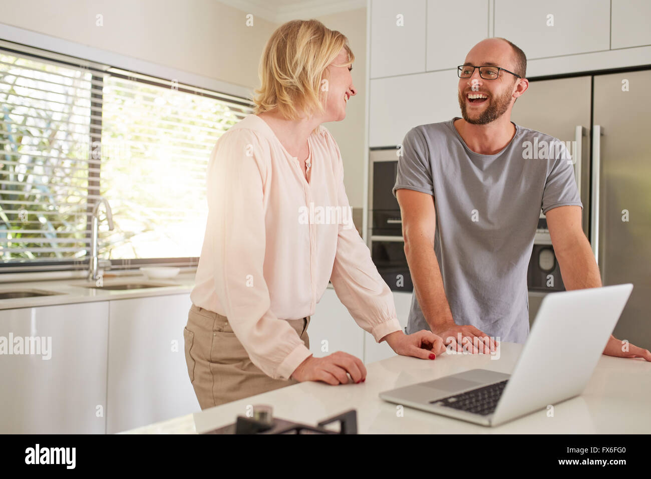 Indoor shot of a happy couple standing at kitchen counter with laptop computer. Man and woman looking at each other and smiling. Stock Photo