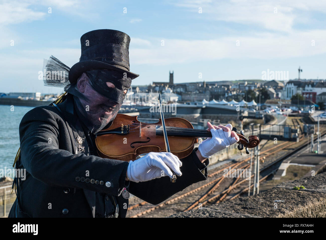 A masked guiser plays the violin with the town of Penzance in the background. Stock Photo