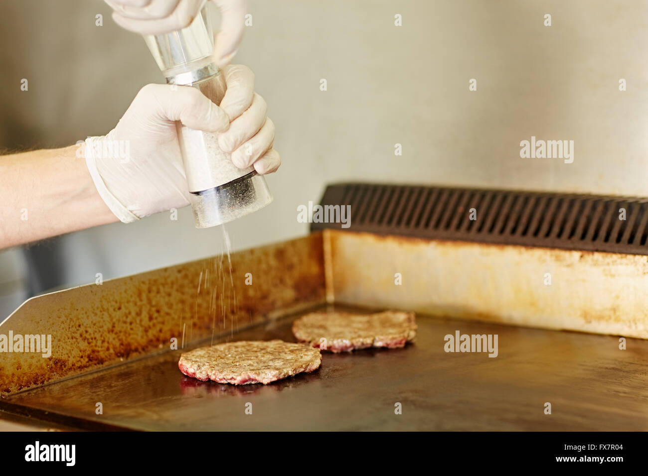Gloved hands grinding salt onto two frying hamburger patties Stock