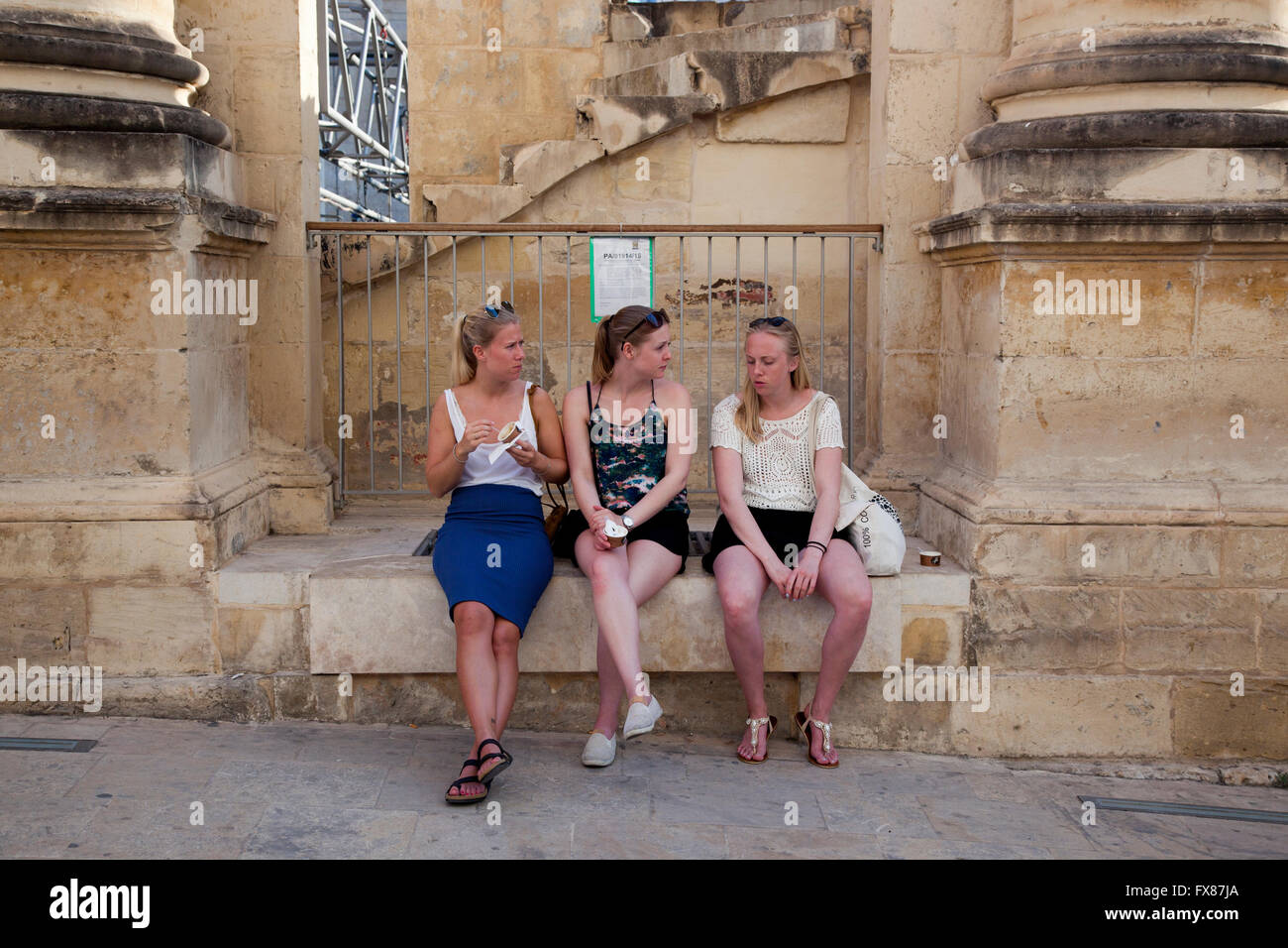 Three tourists take a break from touring in Valletta by sitting outside the ruins of the Royal Opera House. Stock Photo