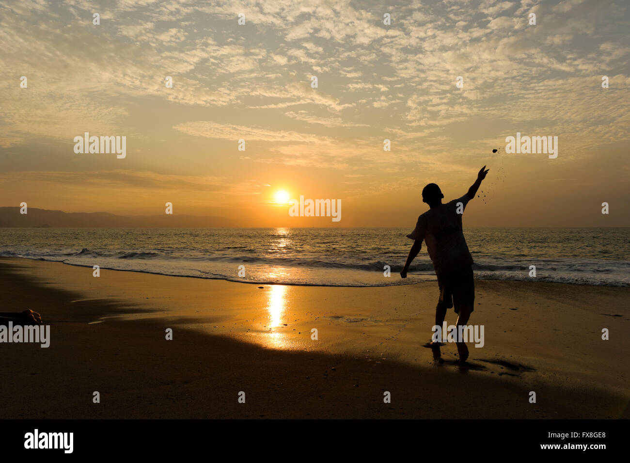 Beach boy is a boy throwing a stone at the the sunset ocean with the sun setting on the horizon. Stock Photo