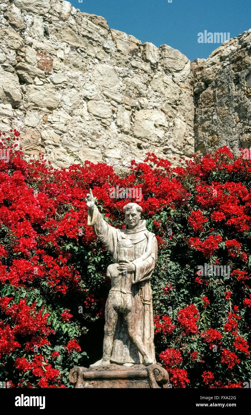 Bright bougainvillea flowers surround a statue of Spanish missionary Junipero Serra embracing a young Indian boy, a 1914 monument by American artist Tole Van Rensaalar in the courtyard of Mission San Juan Capistrano in Orange County, California, USA. The fabled Franciscan friar founded the mission in 1776, the seventh of 21 California missions established up and down the state to expand Spain’s territory and convert the Native Americans to Christianity. Father Serra was canonized as a Catholic saint by Pope Francis in 2015. Stock Photo