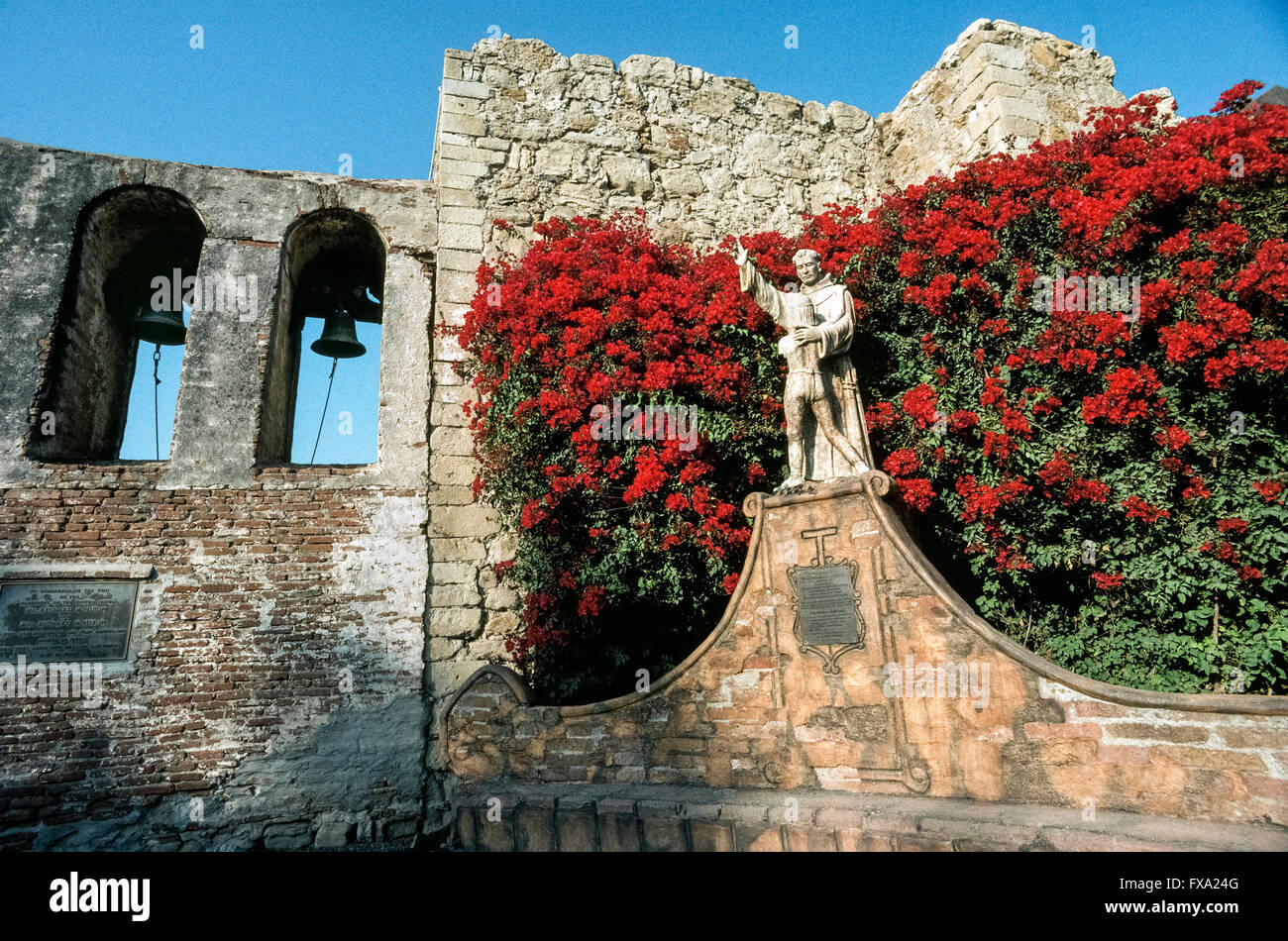 Bright bougainvillea flowers surround a statue of Spanish missionary Junipero Serra embracing a young Indian boy, a 1914 monument by American artist Tole Van Rensaalar in the courtyard of Mission San Juan Capistrano in Orange County, California, USA. The fabled Franciscan friar founded the mission in 1776, the seventh of 21 California missions established up and down the state to expand Spain’s territory and convert the Native Americans to Christianity. Father Serra was canonized as a Catholic saint by Pope Francis in 2015. Stock Photo