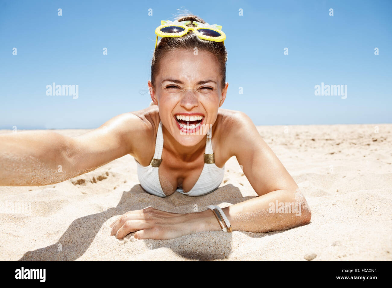 Heading to white sand blue sea paradise. Smiling woman in white swimsuit and funky glasses taking selfies while laying on sandy Stock Photo