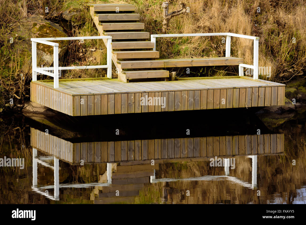Hovering wooden jetty over still and motionless water. White railing and unpainted steps leading up from the jetty. Stock Photo
