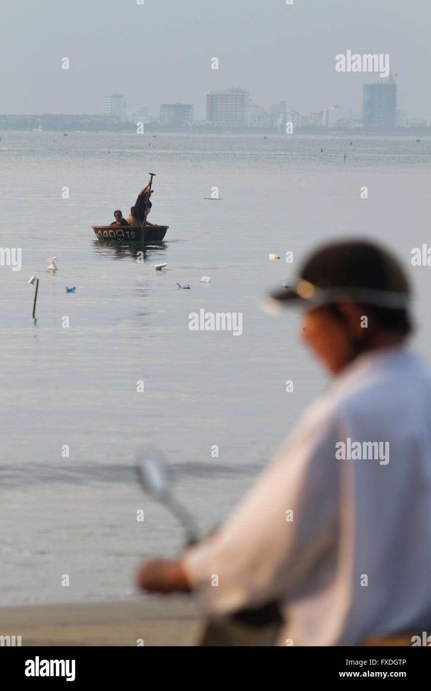 Vietnamese man with motorbike; coracle boat & skyline at Danang beach, Vietnam Stock Photo