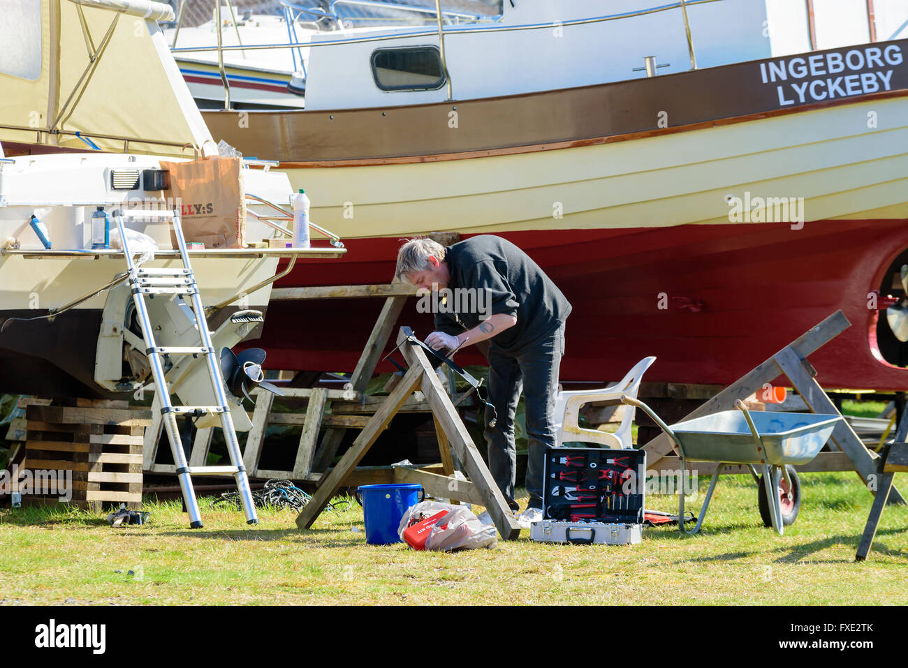 Lyckeby, Sweden - April 7, 2016: Adult male cleaning or repairing a boat detail on a boat yard. Open toolbox beside him. He wear Stock Photo