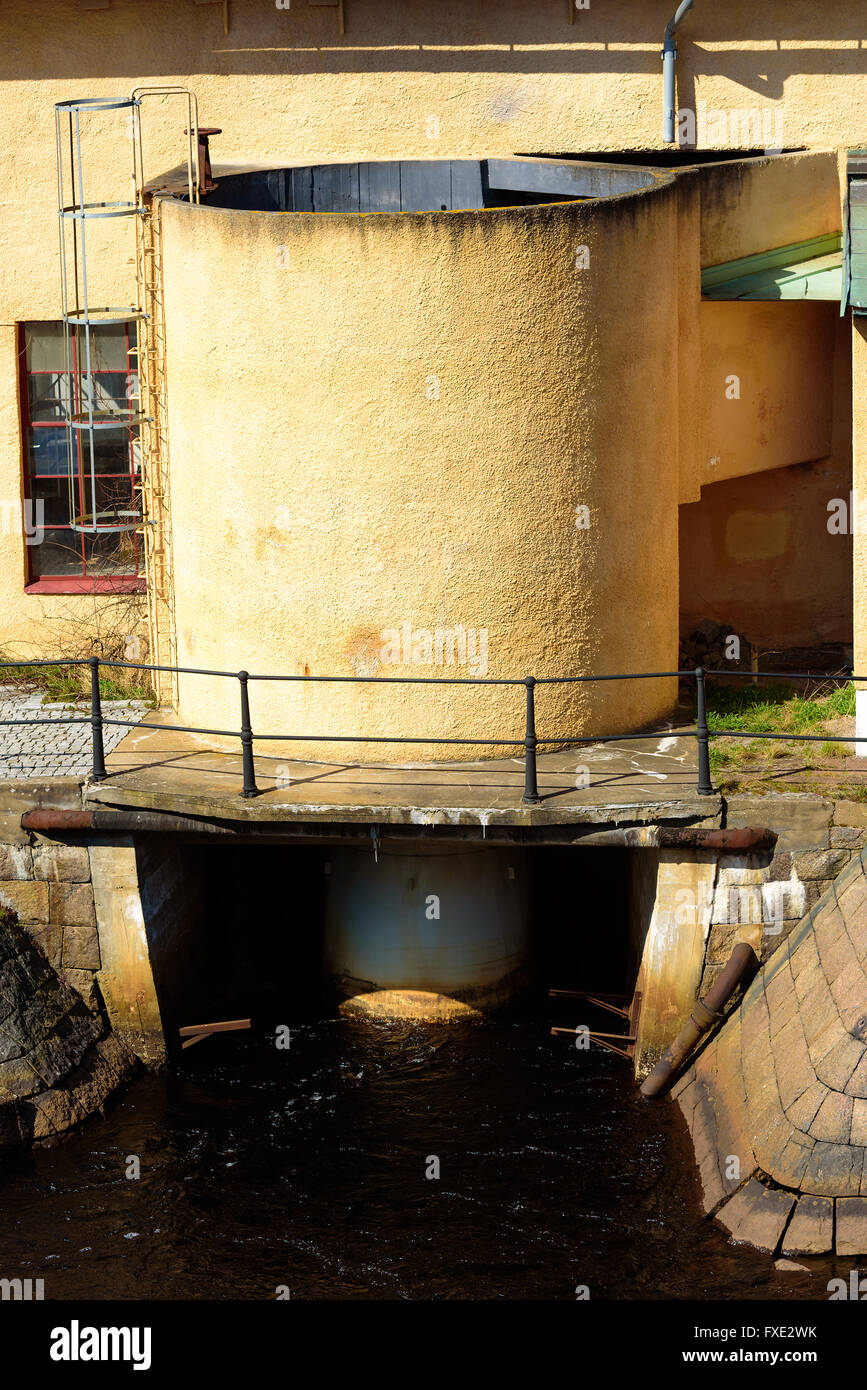 Open water cylinder or silo outside an old mill. Water flows out from underneath and out into the passing river. A ladder leads Stock Photo