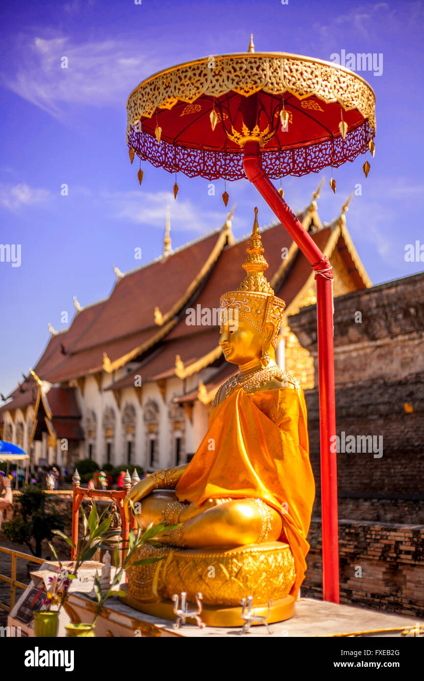 Gold Buddha sitting statue under umbrella with blue sky and temple in